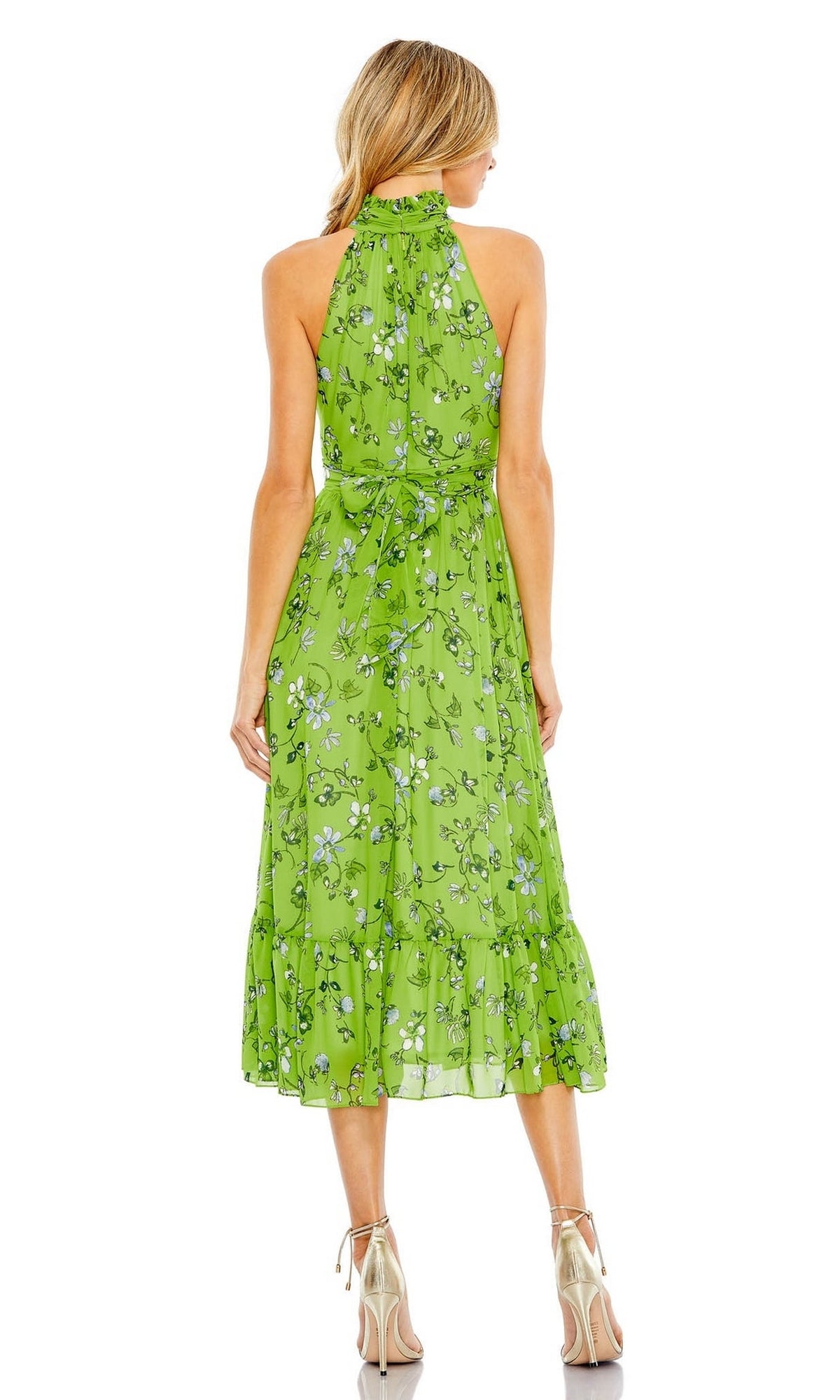 A woman faces away from the camera, wearing the Mac Duggal Short Wedding Guest Dress 55854 in sage green with floral prints, halter-neck and ruffle details, paired with metallic high-heeled sandals.